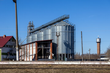 Industrial grain storage silos made of corrugated metal sit near a railway track and a brick tower under a clear blue sky, representing modern agricultural infrastructure and trade © Alina