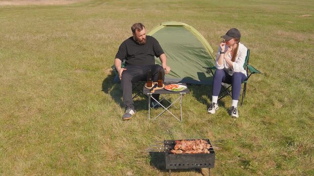 The bearded man gestured and yelled at his bored girlfriend during a tense altercation while camping in a field with a tent, barbecue grill, and food on a small table
