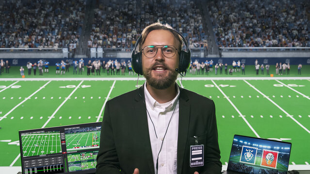 White Male Broadcaster Wearing Glasses, Headset Microphone and Press Pass Speaks to Camera While Covering a Packed Football Arena, Dual Laptops Displaying Game Footage and Club Emblems.