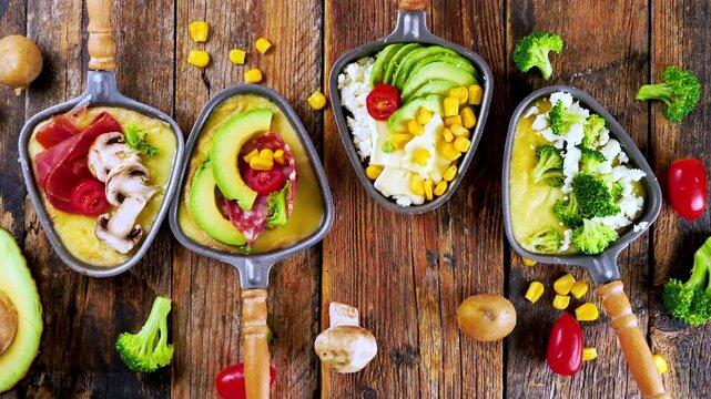 melted raclette cheese in small pans with healthy toppings avocado broccoli corn on rustic wooden background