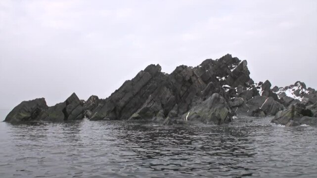Angular Rocky Islet Rising from Calm Water in Cold Coastal Seascape. The frame also shows steep layered black rock formation with sharp ridges, sparse snow and gray water in foreground.