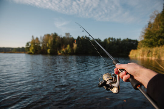 Close up of an angler holding a spinning fishing rod with a reel over a calm river or lake, practicing active lure fishing during a sunny day.
