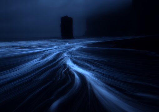Solitary rock pillar standing in dark moody ocean water with glowing blue tidal movement for atmospheric coastal scenes