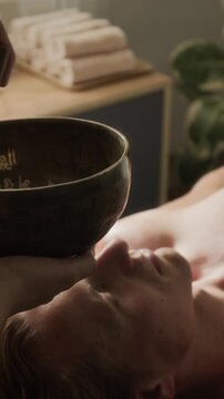 Vertical video of therapist playing Tibetan singing bowl above head of young adult Caucasian man. Clip suitable for holistic wellness marketing, sound therapy and stress relief campaigns
