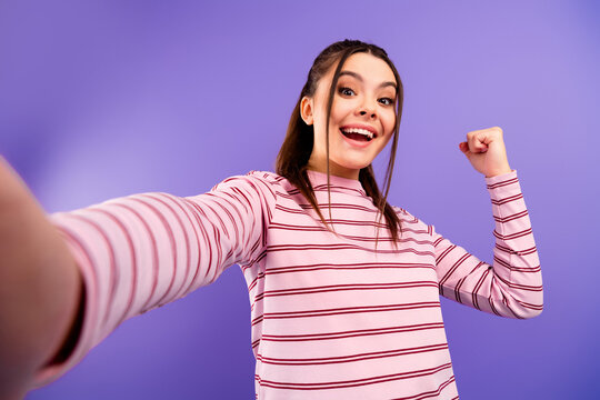 Happy teen girl taking a victorious selfie with arm raised, excited casual portrait on purple background celebrating success and positive mood