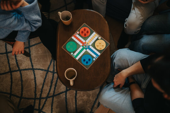 Friends at home play a colorful board game on a small coffee table. Overhead view shows relaxed people gathered for a casual game night with drinks.