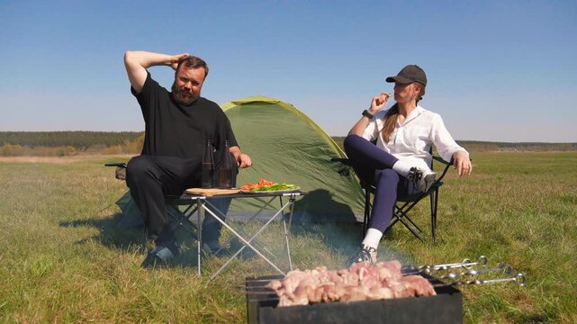 Bearded man and young woman relaxing and talking near a tent in a grassy field, enjoying a barbecue picnic with fresh vegetables and drinks on a folding table during their camping trip