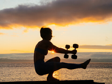 White woman exercising balancing with dumbbells at sunset by water