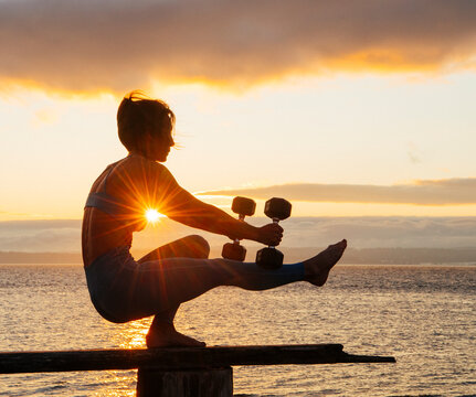 White woman balancing with dumbbells on wooden beam at sunset by water