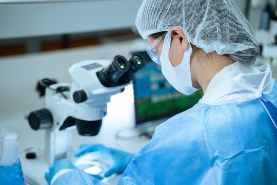 A researcher wearing protective gear focuses on analyzing samples using a stereo microscope in a laboratory, showing dedication and precision during scientific experiments and medical testing.