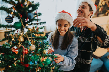 Naklejka premium A young couple decorates a Christmas tree together, both wearing Santa hats and smiling. The festive scene shows them placing ornaments and garlands on the tree in a cozy home setting.