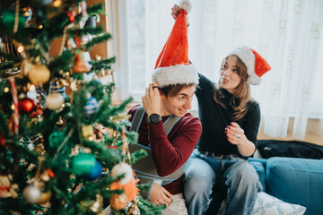 Naklejka premium A young couple wearing Santa hats sit by a decorated Christmas tree and laugh while they decorate. The pair share a playful moment trimming ornaments and enjoying a cozy holiday scene.