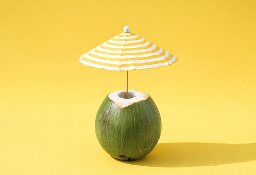 A green coconut topped with a small striped beach umbrella on a bright yellow background, creating a playful tropical summer concept.