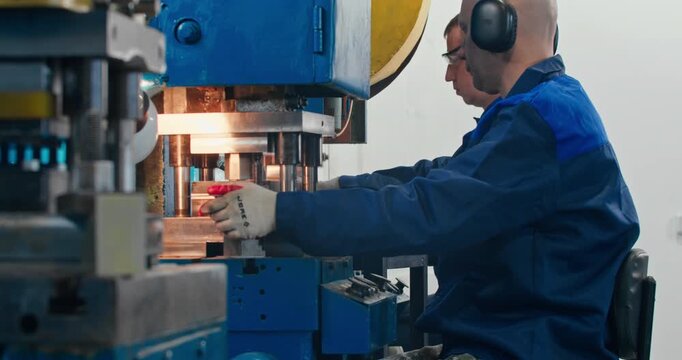 Blue collar factory workers operating stamping machinery during metal parts production. Industrial employment and shop floor work, medium shot