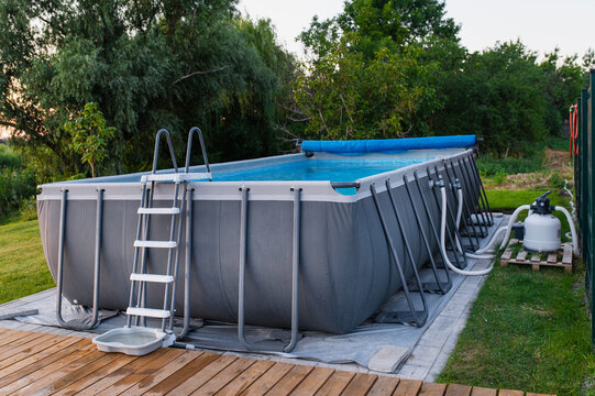 Frame swimming pool with metal ladder stands on wooden deck beside green lawn, equipped with filter and pump for water cleaning, highlighting summer recreation, outdoor living and backyard leisure.