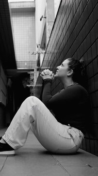 Humble adult female resting on ground with clasped fingers in dark domestic walkway asking for sacred guidance mercy recovery and inner renewal