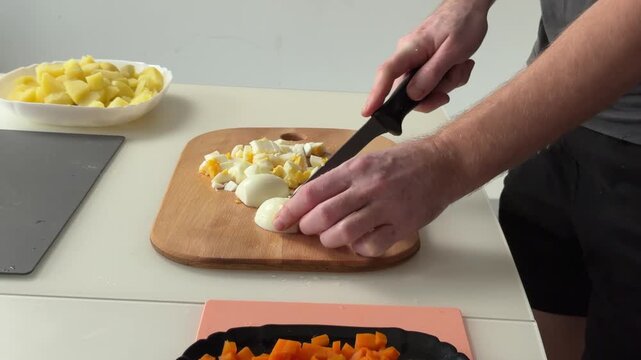 Adult hands chop peeled hard boiled eggs with knife on wooden cutting board, revealing diced egg pieces as cooking prep unfolds on warm countertop light, with hand, knife, cutting, chop detail