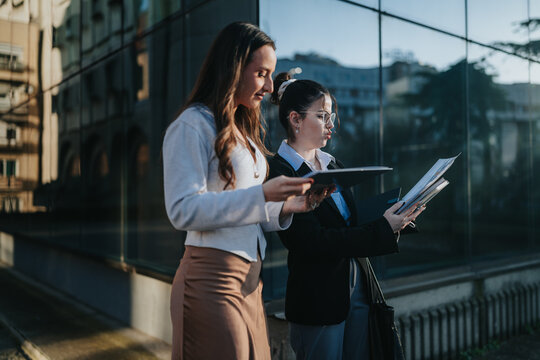 Two businesswomen stand outside an office building reviewing papers and a tablet. They compare documents and discuss work during a brief outdoor meeting.