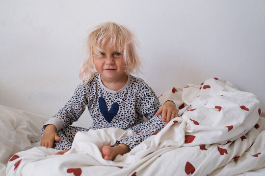 Emotional little girl in pajamas shouting and holding head while sitting on bed