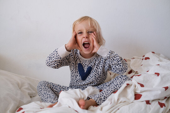 Emotional little girl in pajamas shouting and holding head while sitting on bed