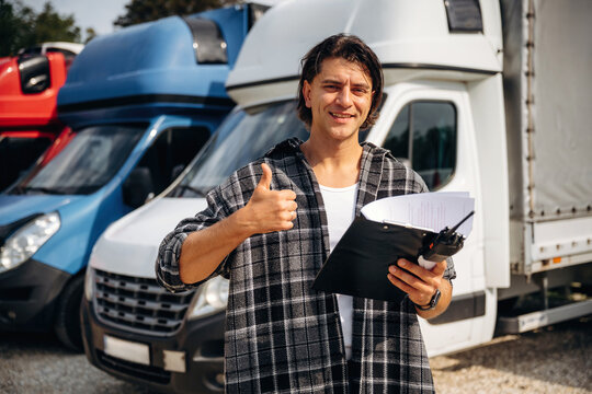 Male logistics manager in checkered shirt holds clipboard and gives thumbs up in front of parked delivery trucks at a transport yard during daytime