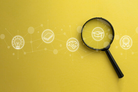 Magnifying glass focusing on a graduation cap icon among educational symbols like brain, book, and lightbulb on yellow background. E-learning, research, and online education discovery concept.