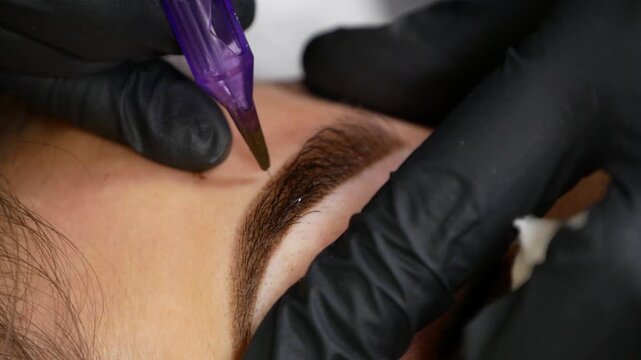 Eyebrow microblading procedure, specialist working on woman's brow with precision tool. Detailed beauty treatment creating defined eyebrow shape and natural-looking appearance