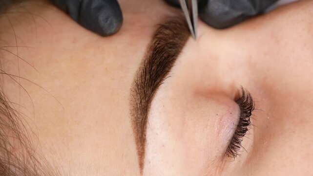 Specialist using tweezers to remove stray eyebrow hairs and refine brow shape after microblading. Precise grooming technique creating clean, defined eyebrow contour