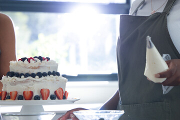 Two-tier round cake is sitting on white pedestal near bright window, topped with assorted berries © WavebreakMediaMicro