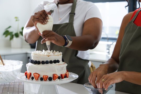 Female co-bakers in olive-green aprons piping with pastry bag on two-tier cake at kitchen bench