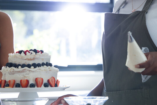 Two-tier round cake is sitting on white pedestal near bright window, topped with assorted berries