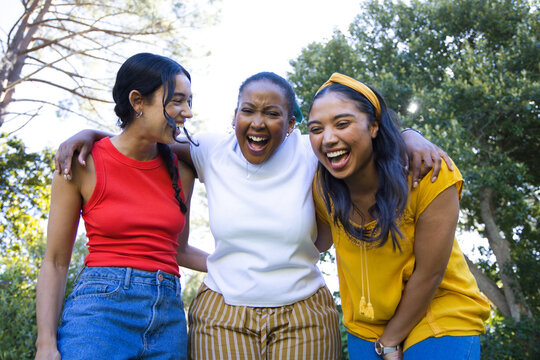 Diverse female friends laughing, leaning to camera in park in red and mustard tops, yellow headband