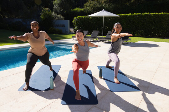 Diverse female friends practicing yoga on dark blue mats by pool, holding lunge with water bottles