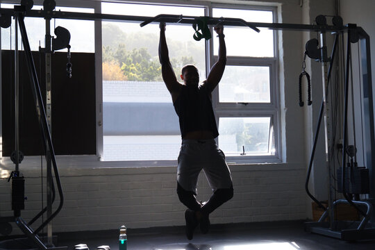 Man performing pullup on horizontal bar at gym wearing workout top and shorts with dumbbells