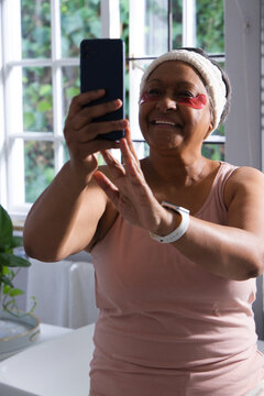Mature African woman holding smartphone, smiling, taking selfie in bathroom with under-eye patches