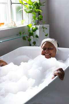 White freestanding bathtub is brimming with foam bubbles in bathroom near frosted window