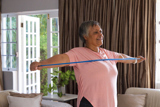 Senior woman wearing pink tee, leggings doing blue resistance-band exercise at home by French doors