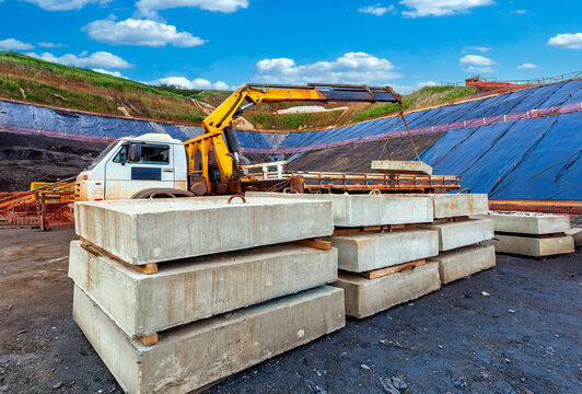 Construction site scene: a truck-mounted crane lifts a precast concrete slab, with stacked concrete units in foreground, and blue landfill liner covering slopes under a cloudy sky.