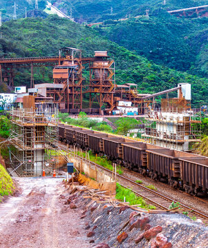Iron ore mining facility in Minas Gerais, Brazil, with a freight train on a railway and bridge infrastructure under construction amidst green mountains.