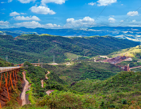 Large iron ore mining operation in Minas Gerais, Brazil, showing vast conveyor belts, power lines, ore stockpiles, and railway transport in a mountainous landscape.