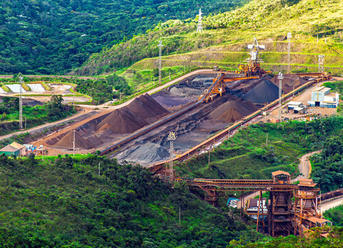 Aerial view of an iron ore mining facility with stockpiles and conveyor belts surrounded by green forests in Minas Gerais, Brazil.