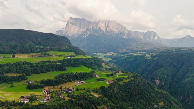 Dramatic Dolomites peaks rising above green valley in South Tyrol, Italy, aerial view of alpine landscape with forest and scattered houses