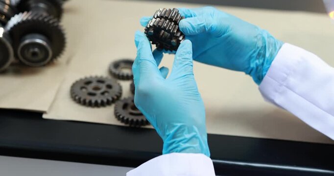 Close-up of a technician wearing blue gloves inspecting a metal gear in a workshop, with machine parts and tools on a workbench.