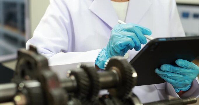 Technician in a white lab coat and blue gloves inspects industrial gears while recording notes on a clipboard in a workshop.
