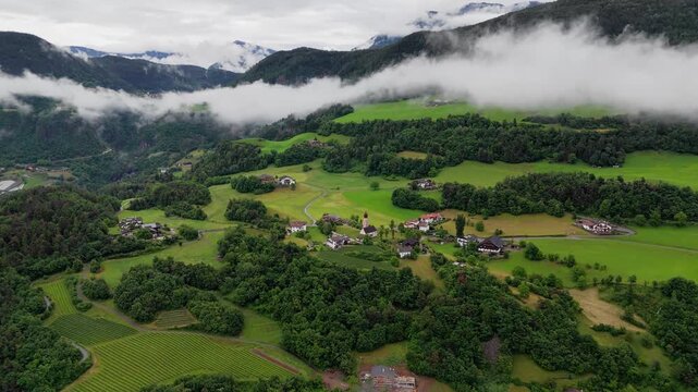 Patchwork fields, woodland and village houses cover green slopes in Dolomites, South Tyrol, Italy. Drone footage