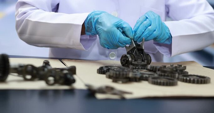 Technician in a white lab coat and blue gloves repairs small metal gears and mechanical parts on a workbench.