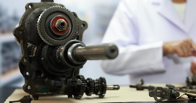 Close-up of disassembled gearbox gears and metal shafts on a workbench, with a mechanic in a white coat repairing machinery in a workshop.