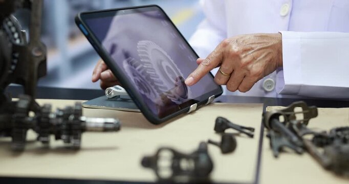 Engineer in a white lab coat uses a tablet to inspect a 3d mechanical part model beside metal components on a workbench in a workshop.