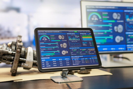 Tablet and desktop monitor display industrial equipment performance dashboards beside a metal machine part on a modern factory workbench.