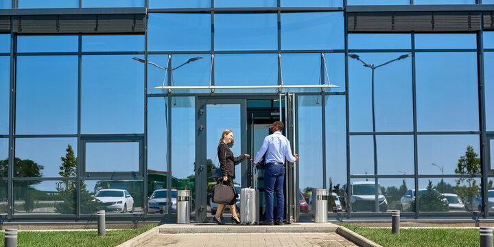 Two corporate travelers discuss directions beside luggage at airport entrance with practical focus. Travel planning, airport navigation and business mobility concept.